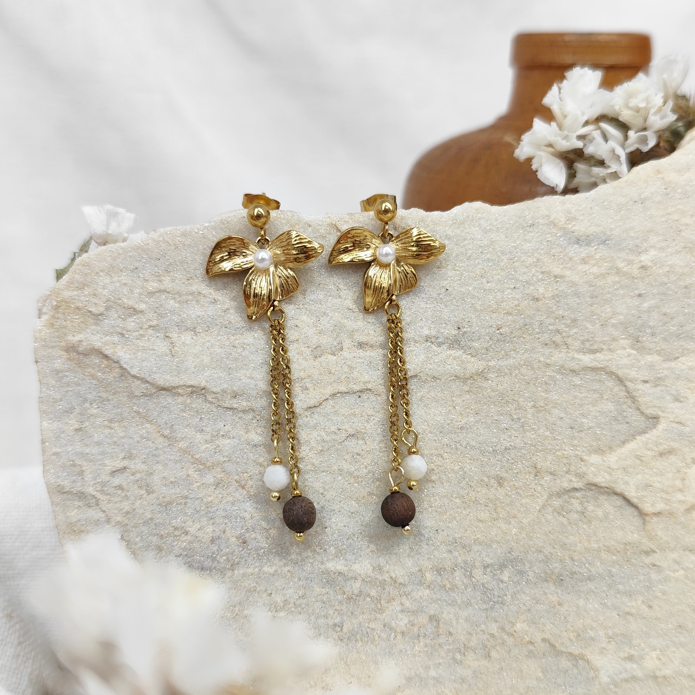 Paar goldene Lilienohrringe mit Kettchen, weißem Naturstein und brauner Nussperle, liegend auf hellem Steinuntergrund