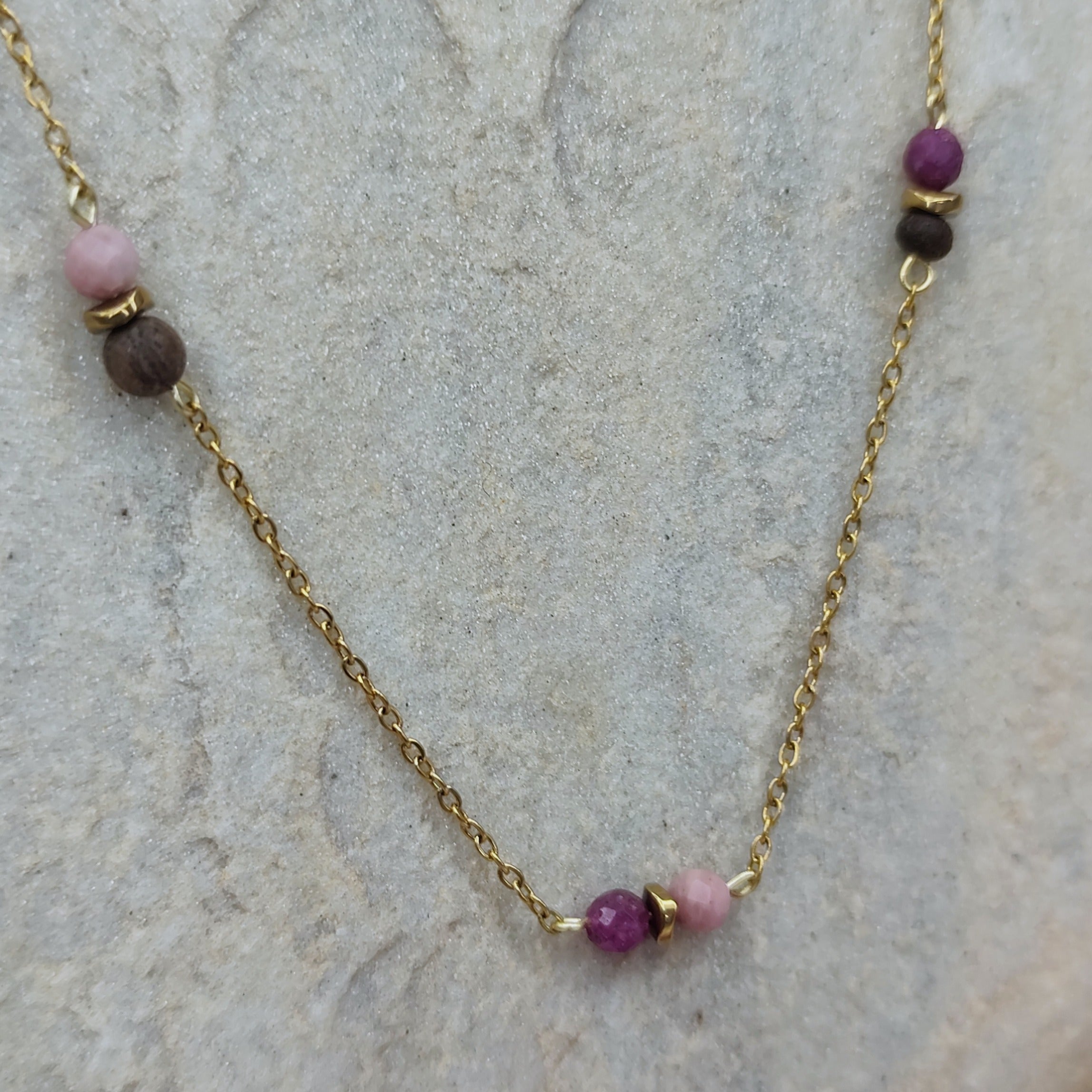 Halskette mit kleinen Nussperlen und farbigen Natursteinen an feiner, goldener Metallkette, liegend auf hellem Stein. Detailaufnahme