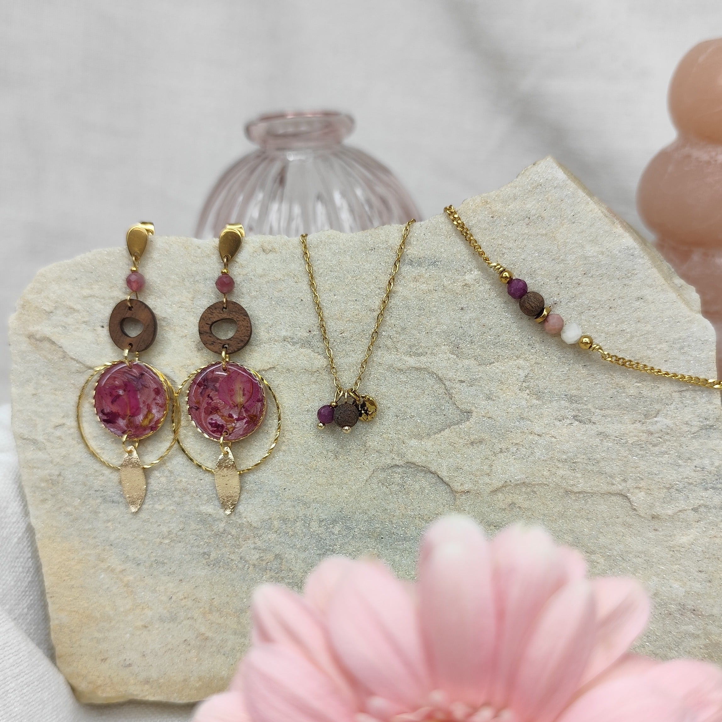 Schmuckset mit Ohrringen, Halskette und Armband aus Nussholz, runden Resin-Elementen mit echten rosa Blüten und goldfarbenen Metallelementen, arrangiert auf hellem Stein mit rosa Blüte im Vordergrund