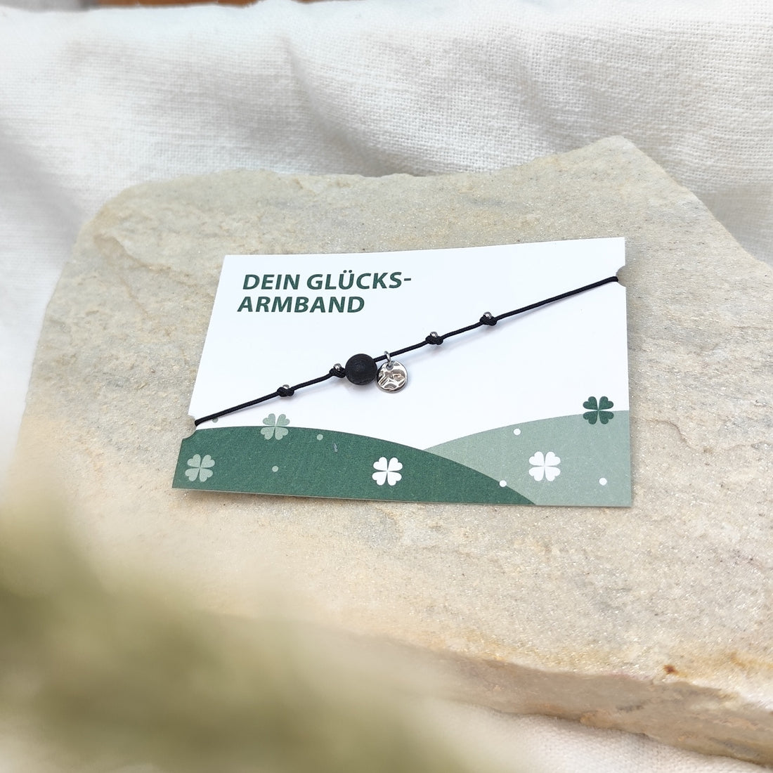 Handgefertigtes Glücksarmband mit Räuchereicheperle und gehämmertem, silbernen Edelstahlplättchen auf naturfarbenem Baumwollband, liegend auf hellem Stein mit Karte