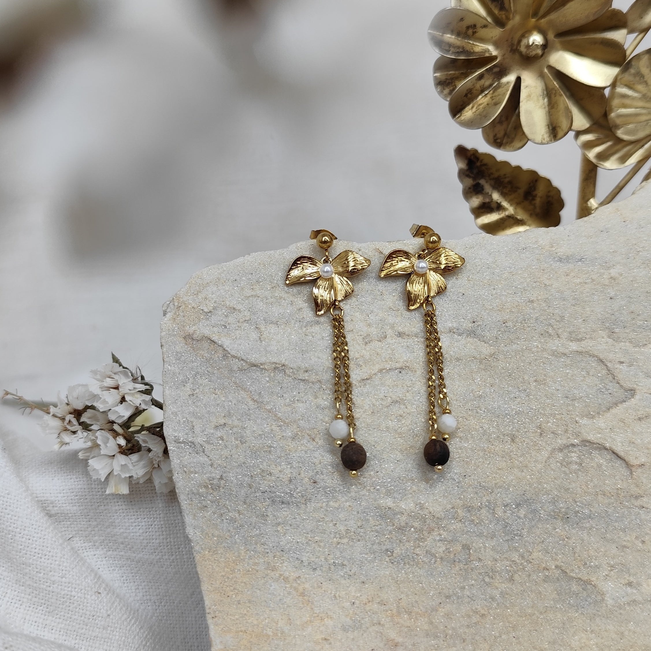 Zwei lange goldfarbene Ohrringe mit Lilienblüte als Stecker, daran ein feines Kettchen mit weißem Naturstein und dunkler Nussperle, präsentiert auf hellem Naturstein