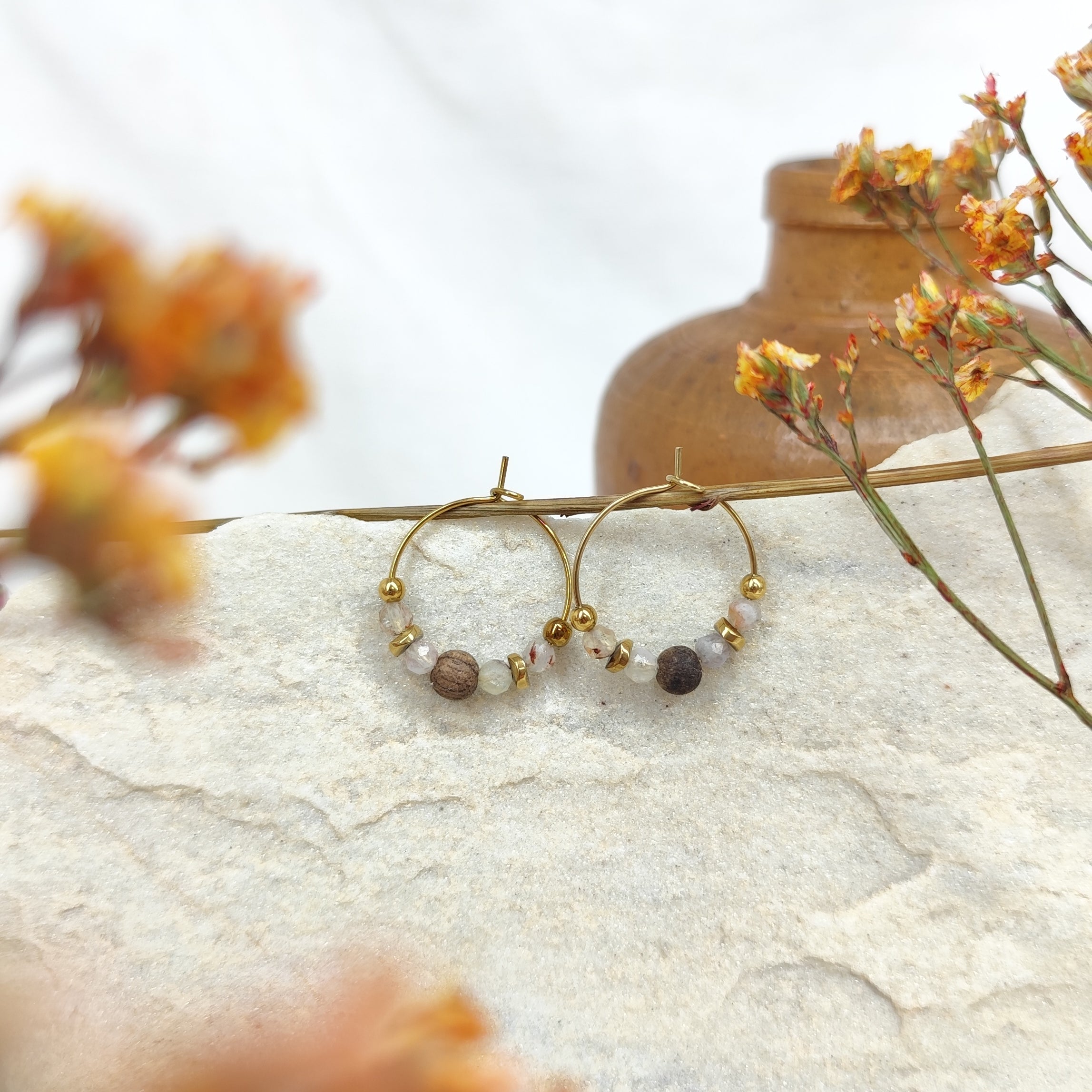 Zwei kleine goldfarbene Kreolen mit je einer hell schimmernden Mondsteinperle und einer braunen Nussperle, liegend auf einem hellen Stein, im Vordergrund unscharfe orangefarbene Blüten