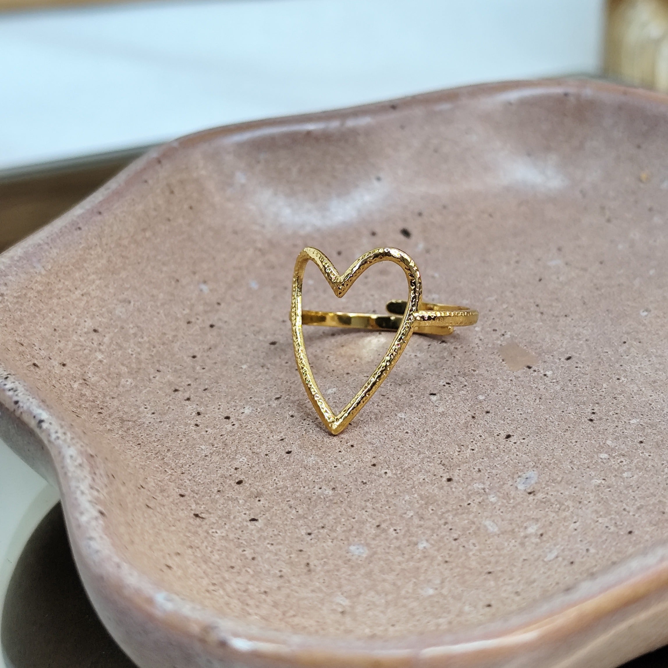 Herzförmiger Ring aus goldenem Edelstahl. Größenverstellbar auf einer grauen Schale mit Spiegel als Untergrund