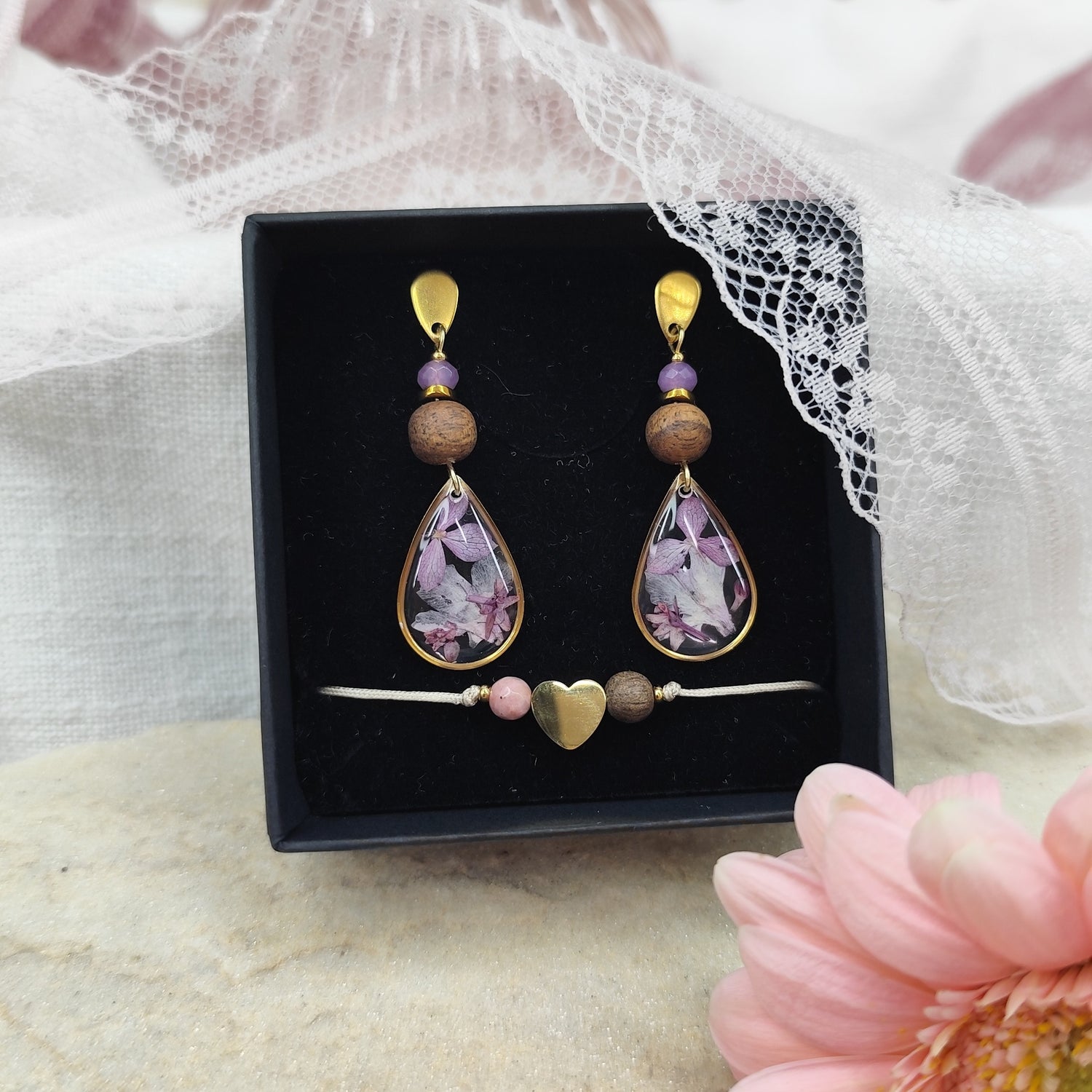 Ohrring mit Naturstein, Nussholz und Resinteil mit echten Blüten in Kombination mit einem Armband aus goldenem Herz und zwei Holzperlen in einer schwarzen Schmuckschachtel. Rosa Blumen und Spitze als Dekoration