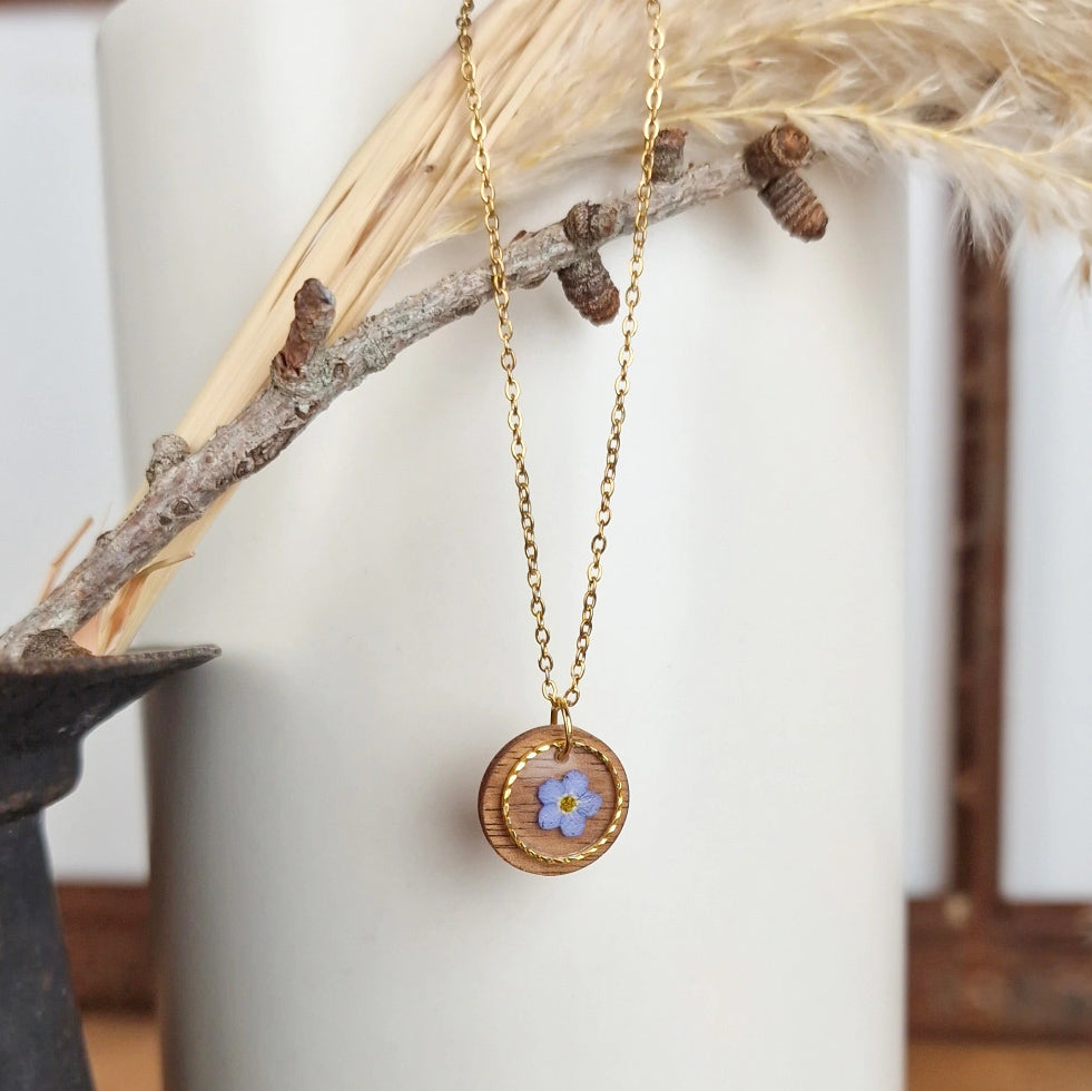 Goldfarbene Edelstahlkette mit rundem Anhänger aus Nussholz, eingefasst mit feinem Metallring und echter blauer Vergissmeinnicht-Blüte im Resin, dekorativ an einem Zweig arrangiert.