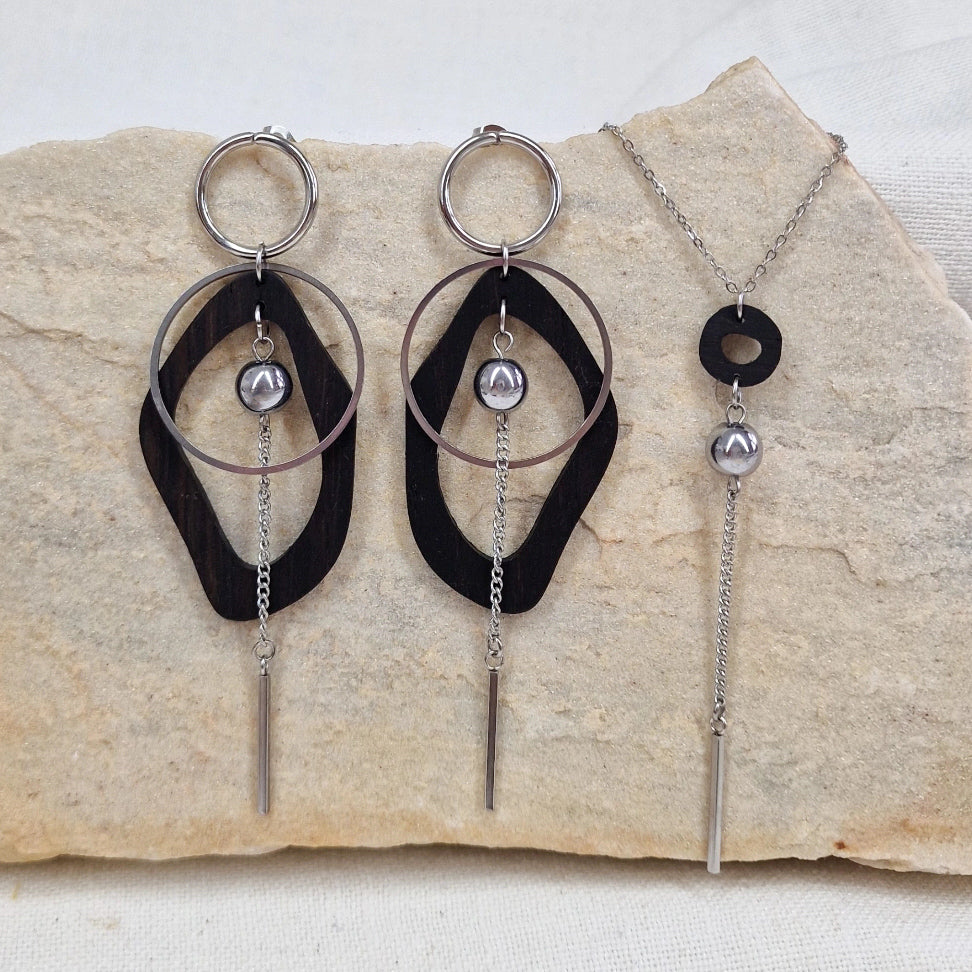 Ohrringe aus Räuchereiche mit silberfarbenen Metallelementen neben einer passenden Halskette, gemeinsam auf hellem Stein liegend