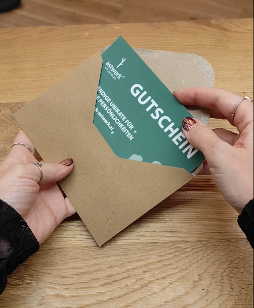 Hände halten einen astwerk® Gutschein in grünem Design und schieben ihn in einen braunen Umschlag auf einem Holztisch.