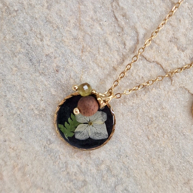 Detailansicht des Anhängers der Halskette ‚Waldlicht‘ aus schwarzem Resin mit echten getrockneten Blüten und Farnen, kombiniert mit Nussholz und Natursteinen