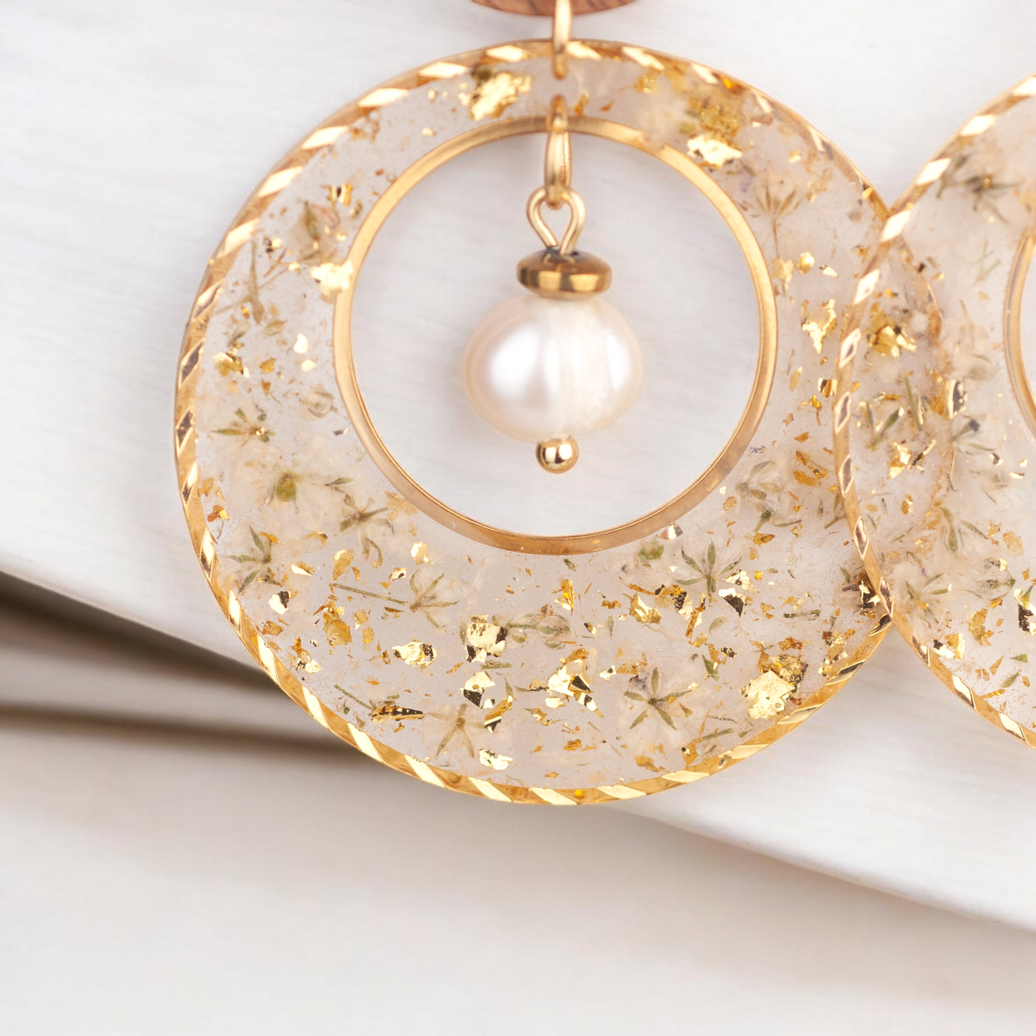 Nahaufnahme der handgefertigten Ohrringe mit transparentem Resin, eingearbeiteten weißen Blüten, Blattgold und mittiger Süßwasserzuchtperle.