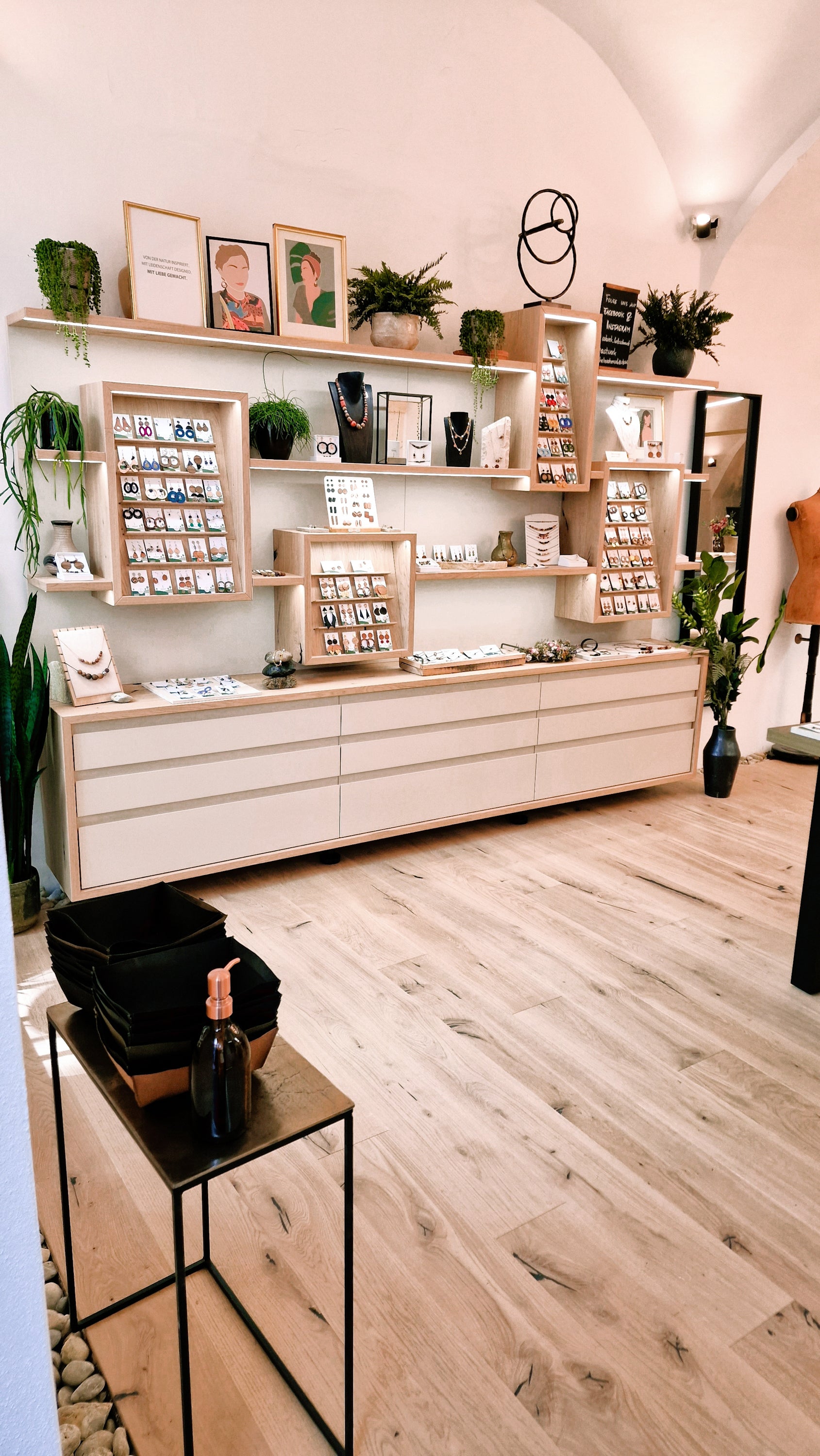 Innenansicht des Astwerk Shops in Graz mit hellem Holzboden, weißer Wand mit Regalsystem aus Holz, präsentiertem handgefertigtem Holzschmuck in Vitrinen und Rahmen sowie Pflanzen und Dekorationselementen im Raum.