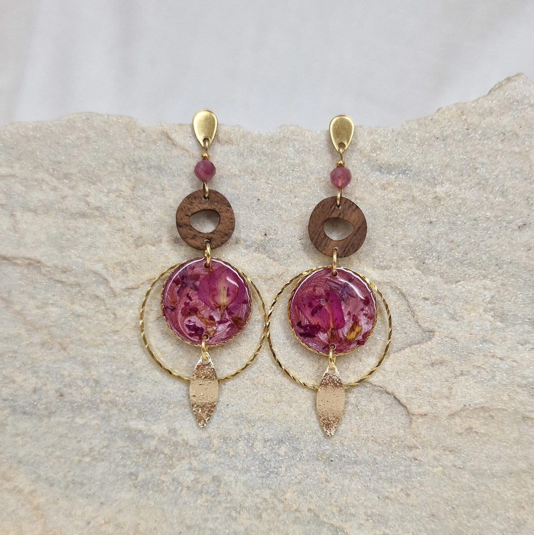 Paar Ohrringe aus goldfarbenem Edelstahl mit Nussholzscheiben und runden Resin-Elementen mit echten rosa Blüten, liegend auf hellem Stein