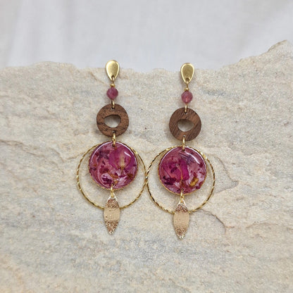 Paar Ohrringe aus goldfarbenem Edelstahl mit Nussholzscheiben und runden Resin-Elementen mit echten rosa Blüten, liegend auf hellem Stein