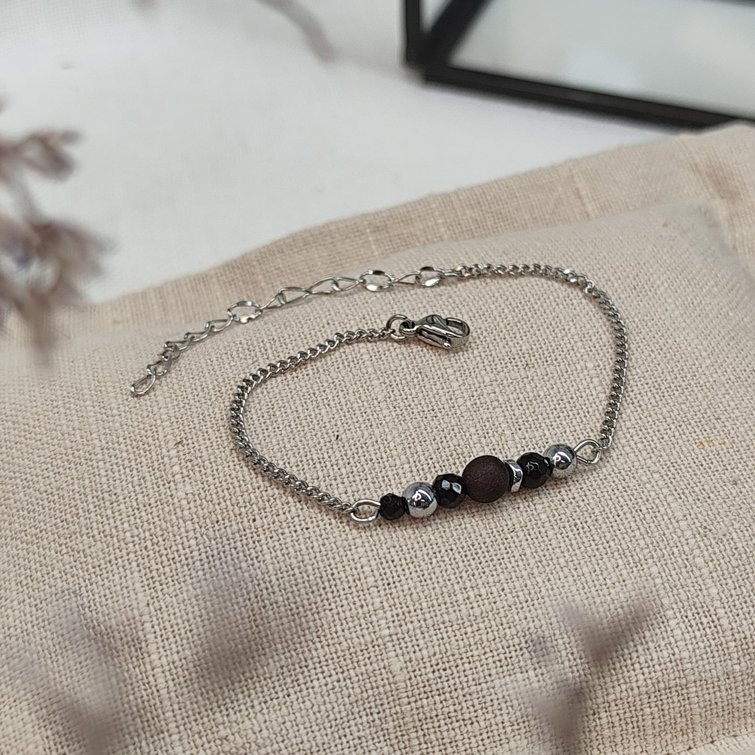 Armband aus Räuchereiche mit Holzperlen und silberfarbener Kette auf einem braunen Webare-Kissen, im Hintergrund eine Ecke Glaskasten und Strauch im Vordergrund