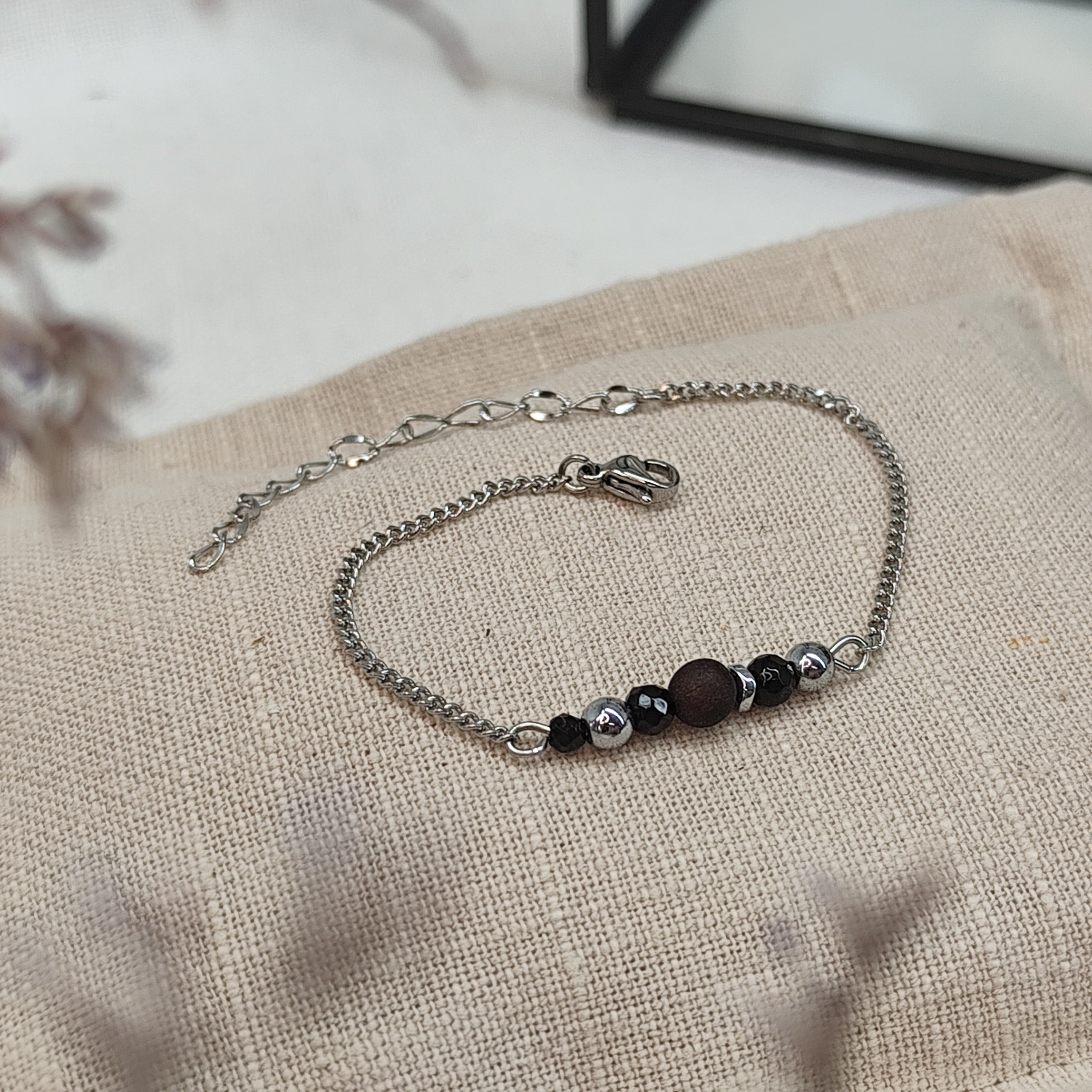 Armband aus Räuchereiche mit Holzperlen und silberfarbener Kette auf einem braunen Webare-Kissen, im Hintergrund eine Ecke Glaskasten und Strauch im Vordergrund