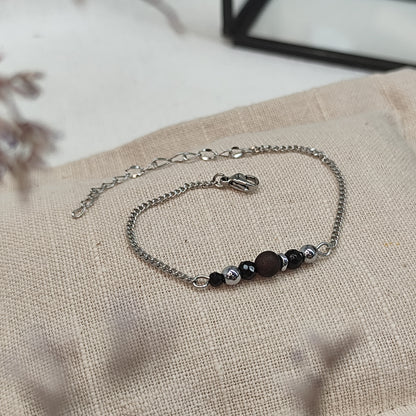 Armband aus Räuchereiche mit Holzperlen und silberfarbener Kette auf einem braunen Webare-Kissen, im Hintergrund eine Ecke Glaskasten und Strauch im Vordergrund