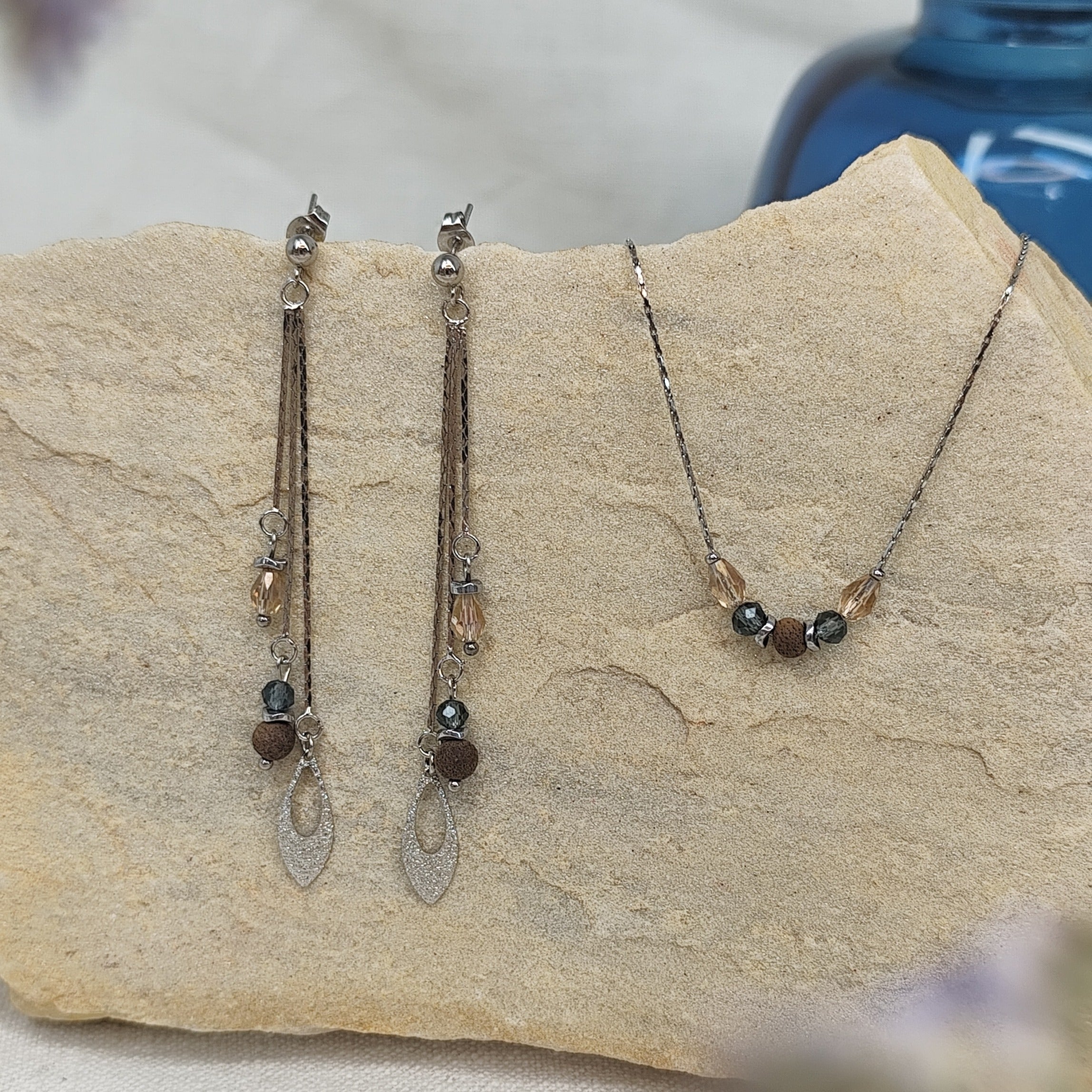 Schmuckset mit Ohrringen und Halskette aus Nussholzperlen, geschliffenen Glaselementen in Blau und Beige sowie Hämatit-Zwischenteilen, liegend auf hellem Stein