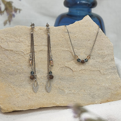 Schmuckset mit Ohrringen und Halskette aus Nussholzperlen, geschliffenen Glaselementen in Blau und Beige sowie Hämatit-Zwischenteilen, liegend auf hellem Stein