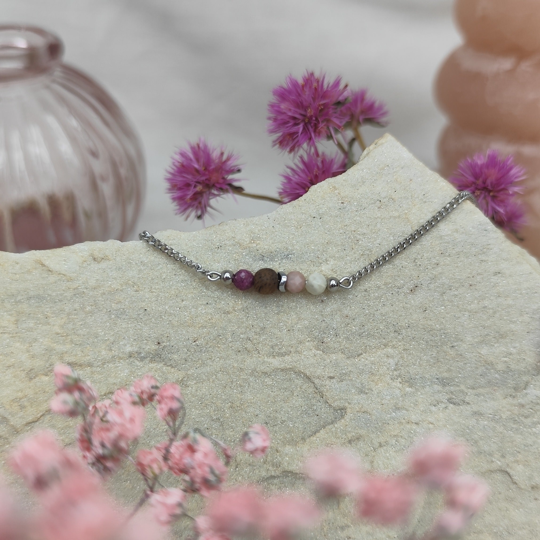 Armketterl mit handgefertigter Nussholzperle, Natursteinen in Rosé- und Violetttönen, arrangiert auf hellem Stein mit rosa Blüten im Hintergrund
