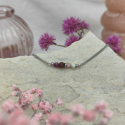 Armketterl mit handgefertigter Nussholzperle, Natursteinen in Rosé- und Violetttönen, arrangiert auf hellem Stein mit rosa Blüten im Hintergrund