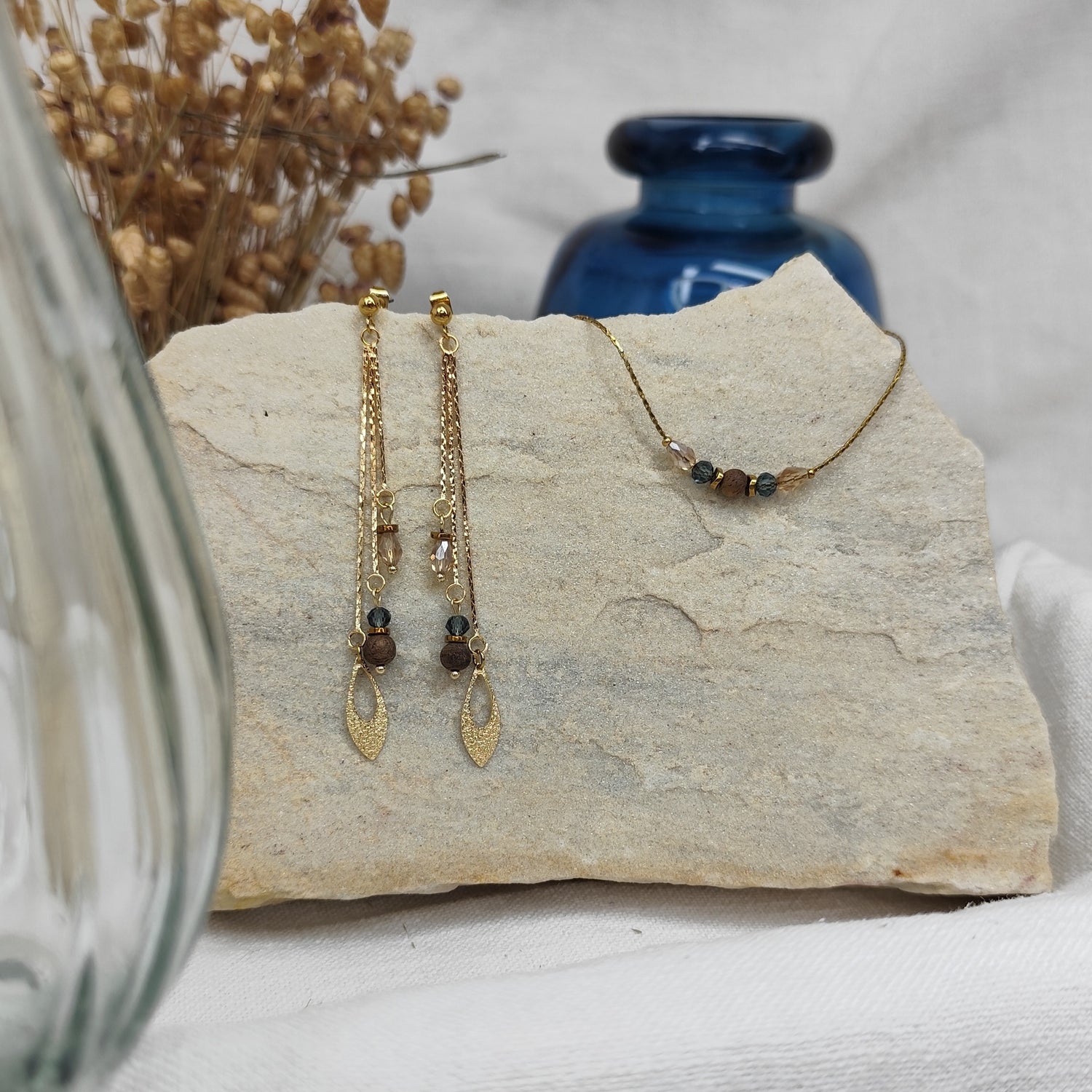 Schmuckset mit Ohrringen und Halskette aus Nussholzperlen, geschliffenen Glaselementen in Blau und Beige sowie Hämatit-Zwischenteilen, liegend auf hellem Stein