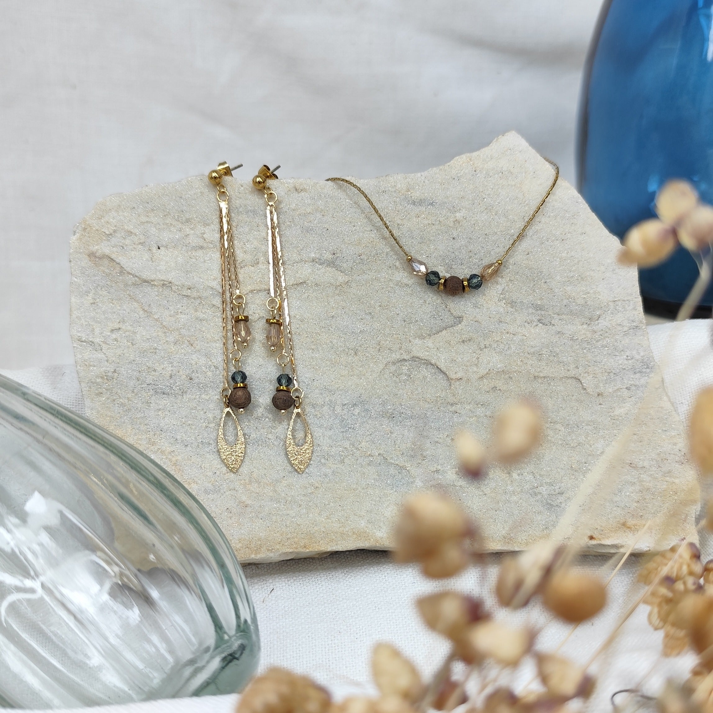 Schmuckset mit Ohrringen und Halskette aus Nussholzperlen, geschliffenen Glaselementen in Blau und Beige sowie Hämatit-Zwischenteilen, liegend auf hellem Stein