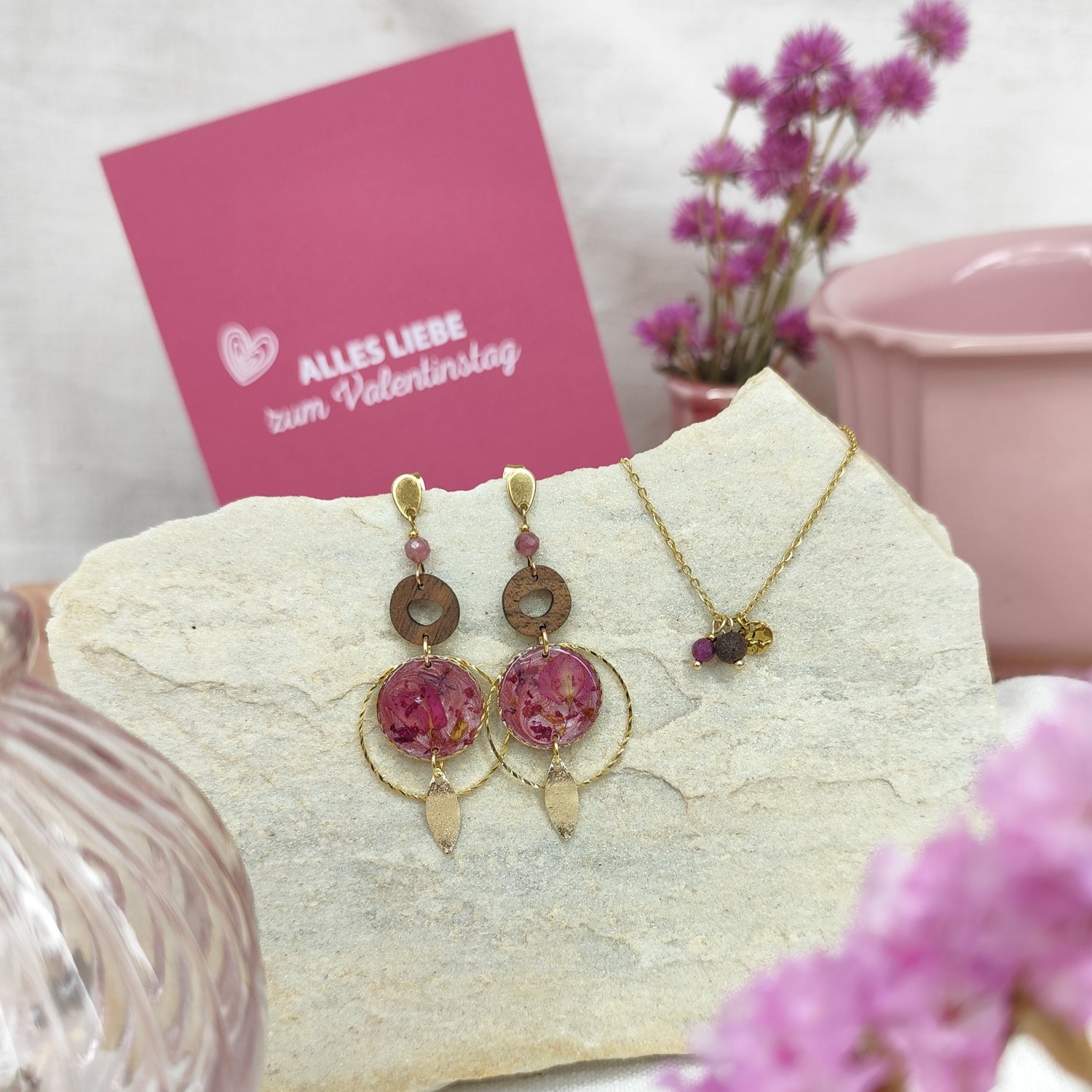 Ohrringe und Halskette aus Nussholz mit Natursteinen und Resin-Elementen mit echten Blüten, gemeinsam auf hellem Stein arrangiert. Im Hintergrund ist eine rosa Karte mit der Aufschrift &