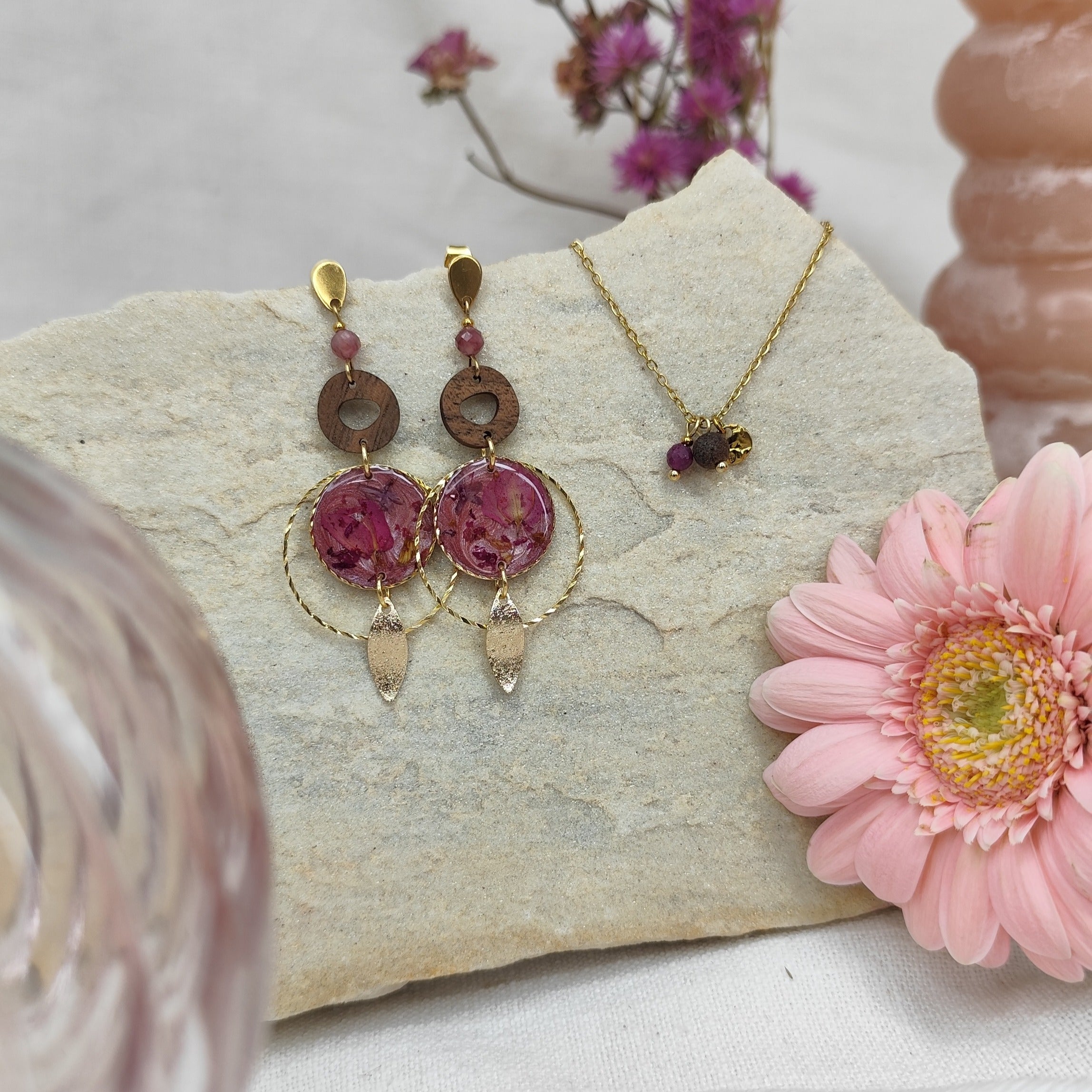Schmuckset mit Ohrringen und Halskette mit Nussholz, Natursteinen und Resin-Elementen mit echten Blumen, liegend auf hellem Stein arrangiert. Im Hinter- und Vordergrund sind rosa Blumen und Vasen als Dekoration