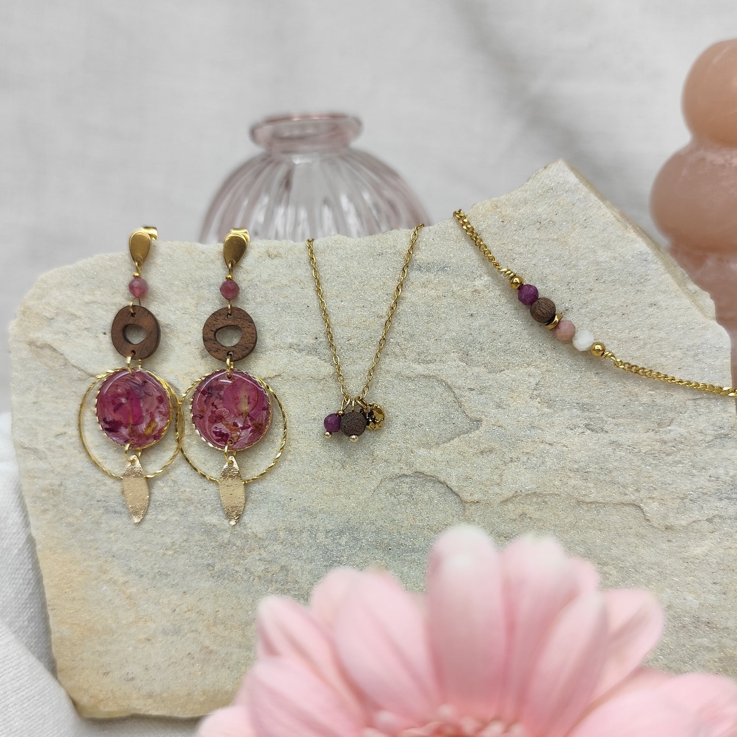 Schmuckset mit Ohrringen, Halskette und Armband aus Nussholz, runden Resin-Elementen mit echten rosa Blüten und goldfarbenen Metallelementen, arrangiert auf hellem Stein mit rosa Blüte im Vordergrund