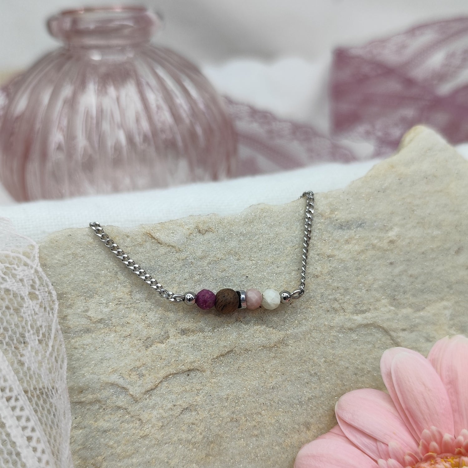 Armketterl mit handgefertigter Nussholzperle, Natursteinen in Rosé- und Violetttönen, arrangiert auf hellem Stein mit rosa Blüten im Hintergrund