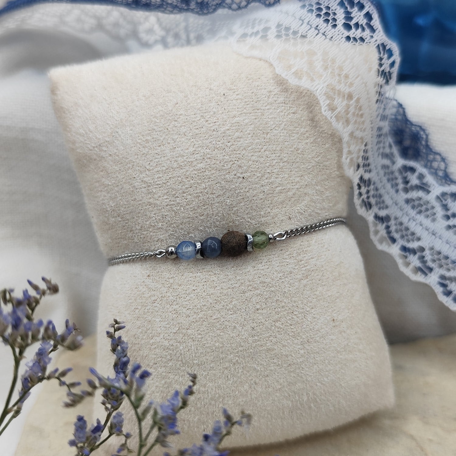 Armband mit blauen und grünen Natursteinen, zentraler Nussholzkugel und Hämatit-Zwischenteilen an feiner Metallkette, liegend auf hellem Stoffpolster