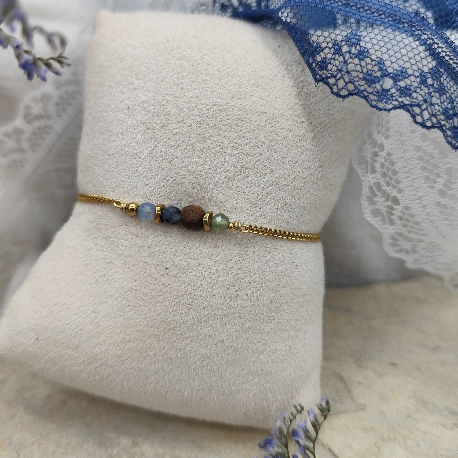 Armband mit blauen und grünen Natursteinen, zentraler Nussholzkugel und Hämatit-Zwischenteilen an feiner Metallkette, liegend auf hellem Stoffpolster