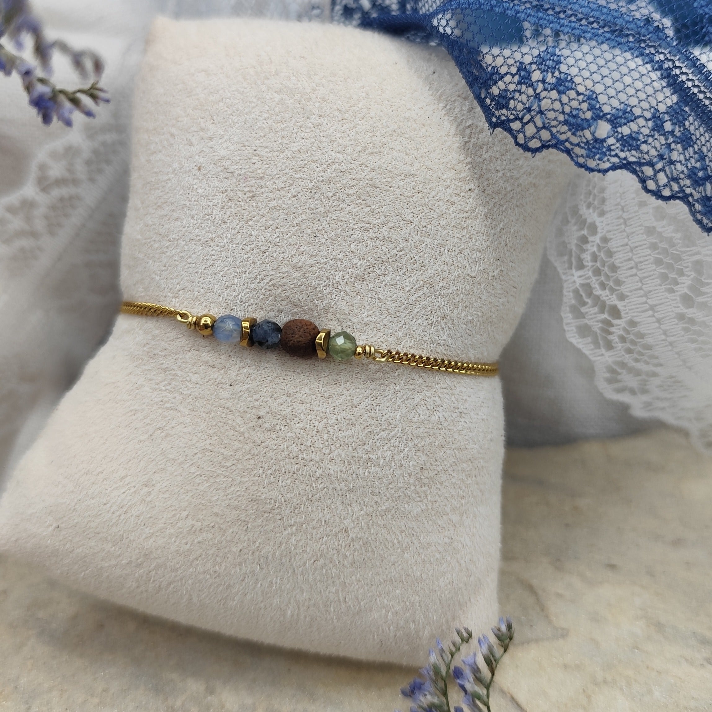 Armband mit blauen und grünen Natursteinen, zentraler Nussholzkugel und Hämatit-Zwischenteilen an feiner Metallkette, liegend auf hellem Stoffpolster