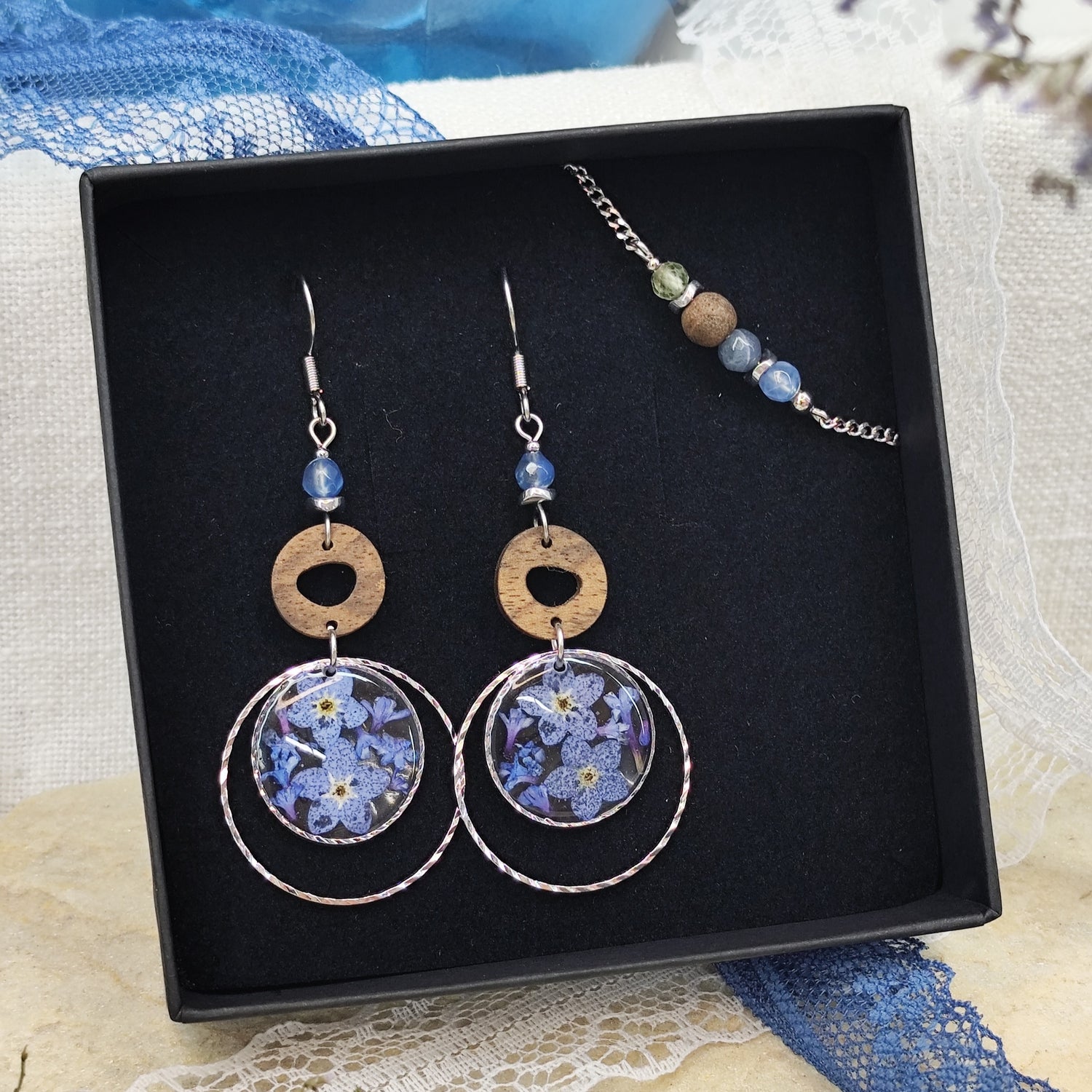 Silberne Ohrringe und Armband aus Nussholz mit runden Resin-Elementen und echten Vergissmeinnicht-Blüten, gemeinsam arrangiert in einer schwarzen Samtschachtel.