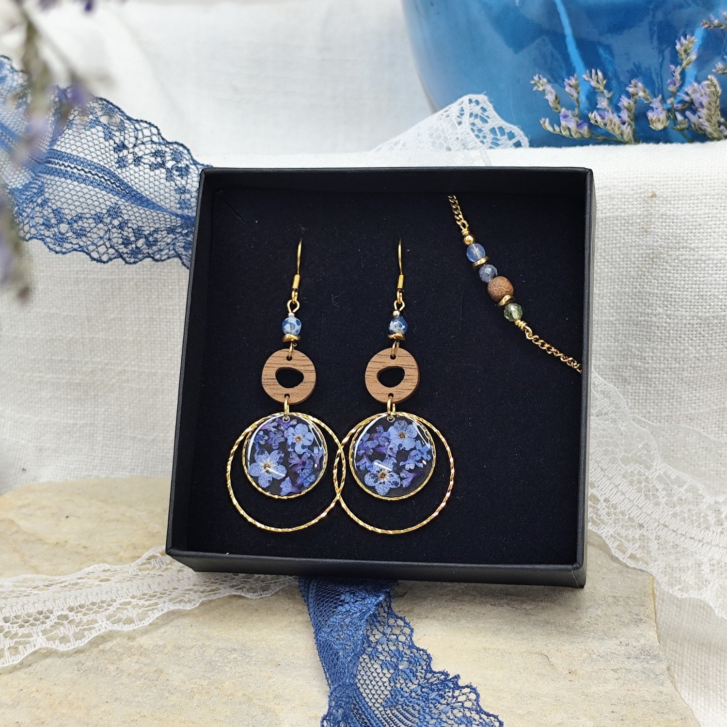 Ohrringe und Armband aus Nussholz mit runden Resin-Elementen und echten Vergissmeinnicht-Blüten, gemeinsam arrangiert in einer schwarzen Samtschachtel. Gold