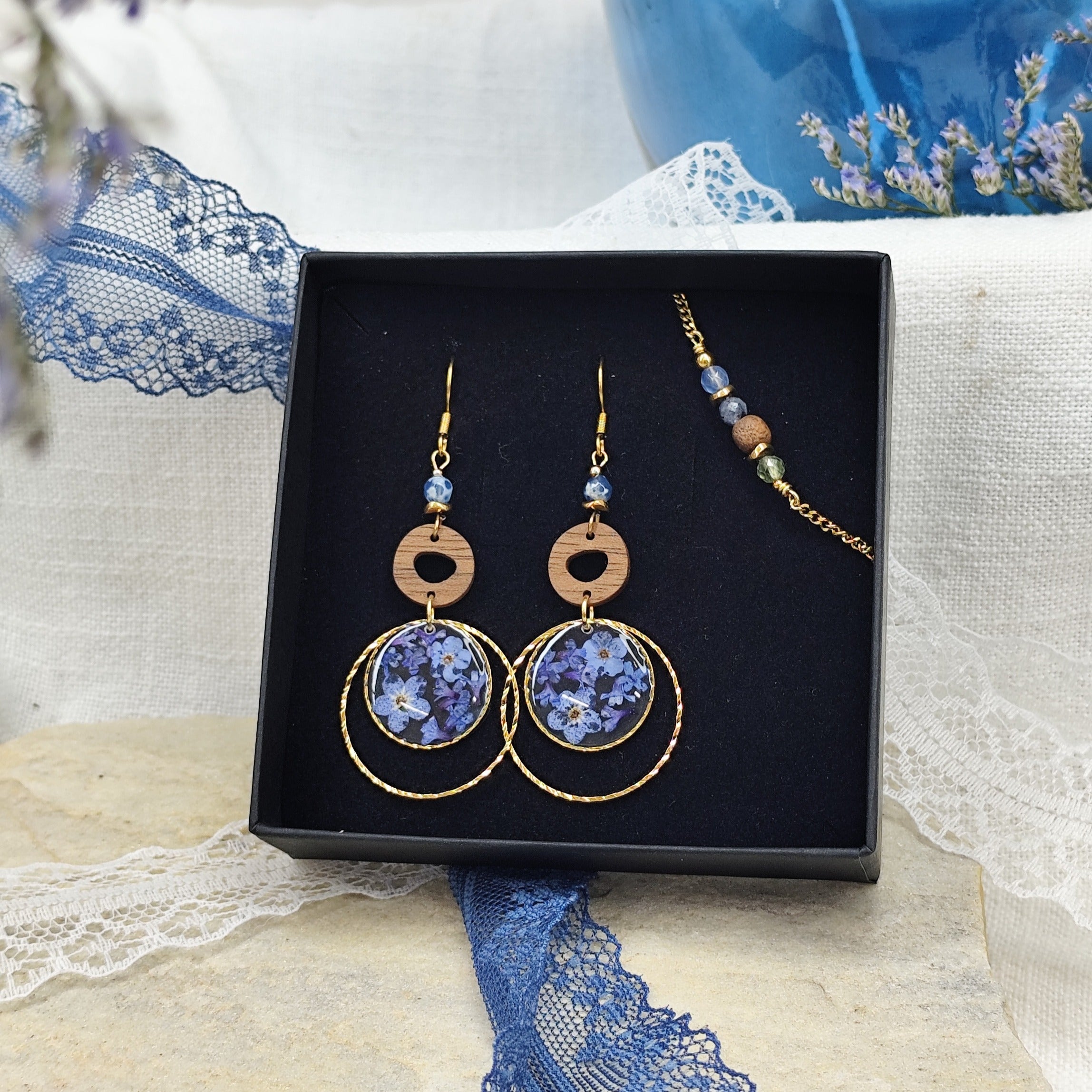 Ohrringe und Armband aus Nussholz mit runden Resin-Elementen und echten Vergissmeinnicht-Blüten, gemeinsam arrangiert in einer schwarzen Samtschachtel. Gold