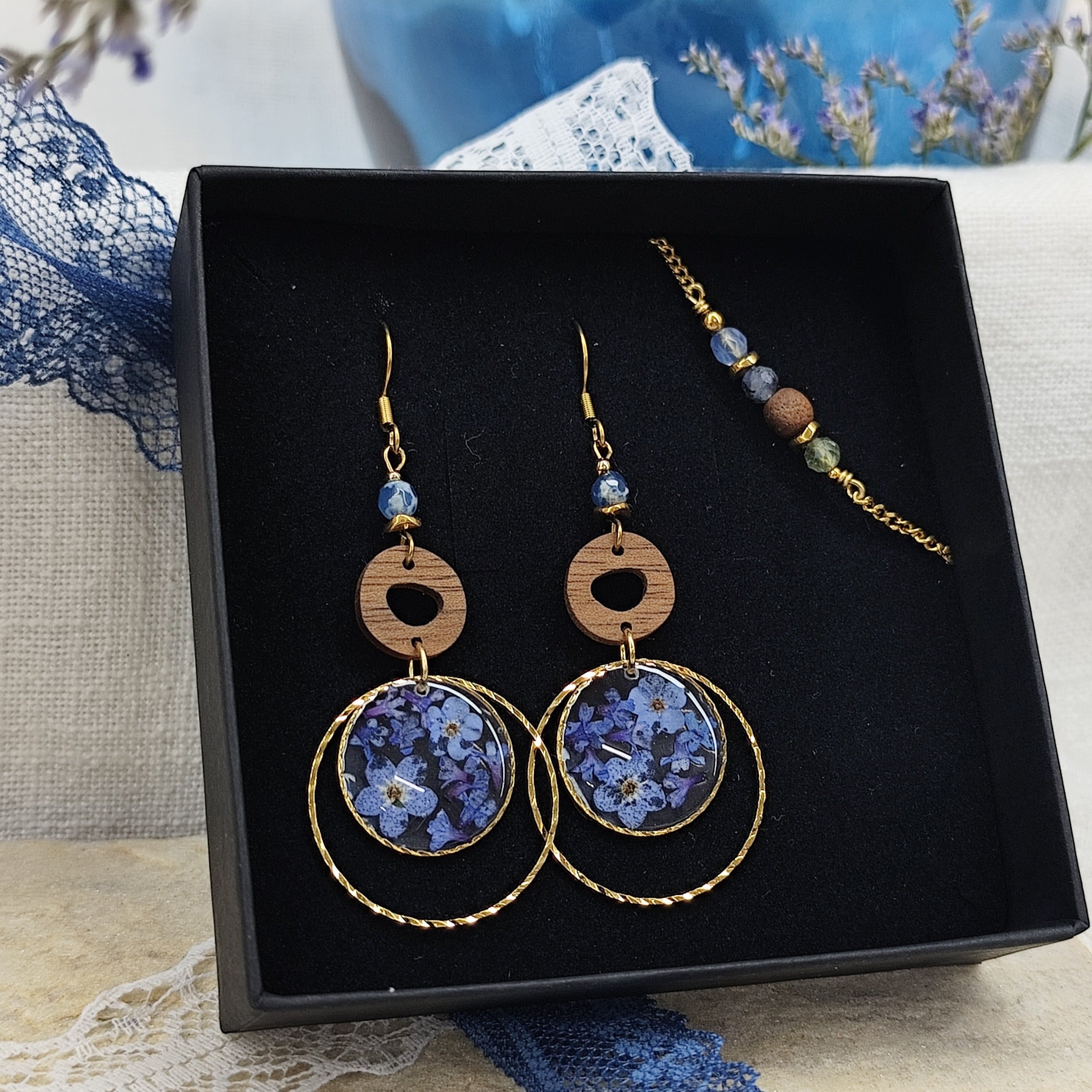 Goldene Ohrringe und Armband aus Nussholz mit runden Resin-Elementen und echten Vergissmeinnicht-Blüten, gemeinsam arrangiert in einer schwarzen Samtschachtel