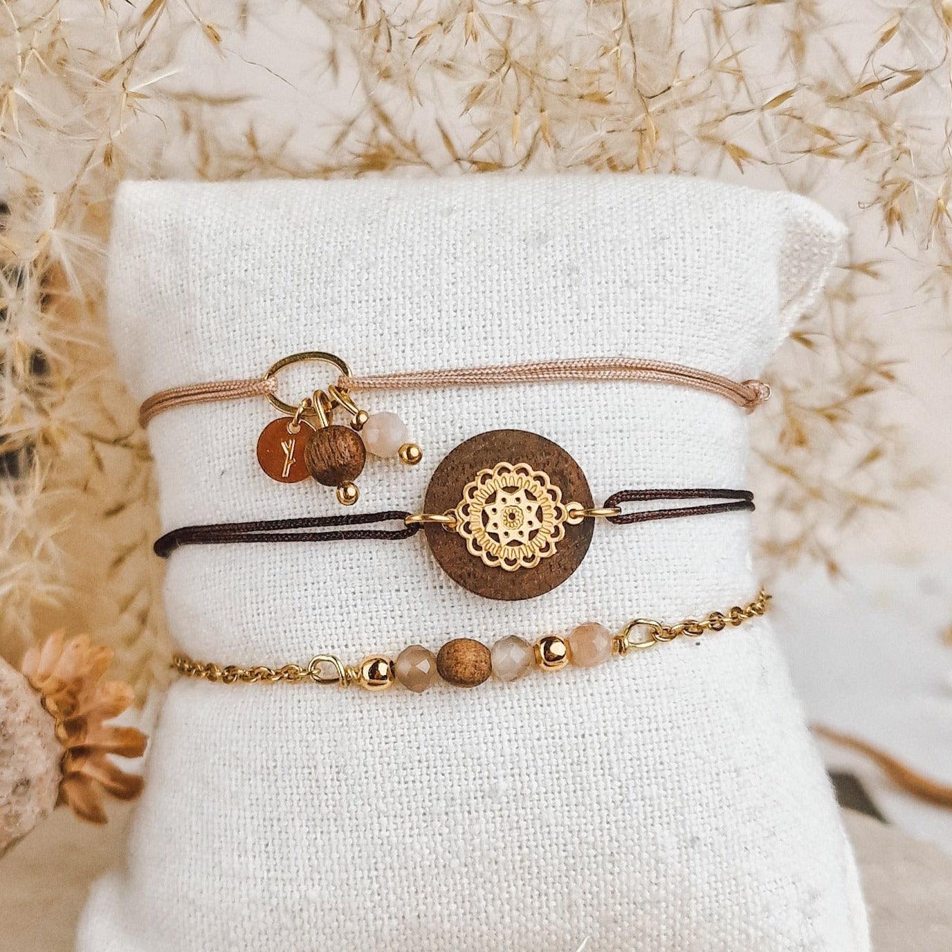 Drei handgefertigte Armbänder mit Nussholz und Mondstein auf einem hellen Baumwollkissen, getrocknete Blumen im Hintergrund.