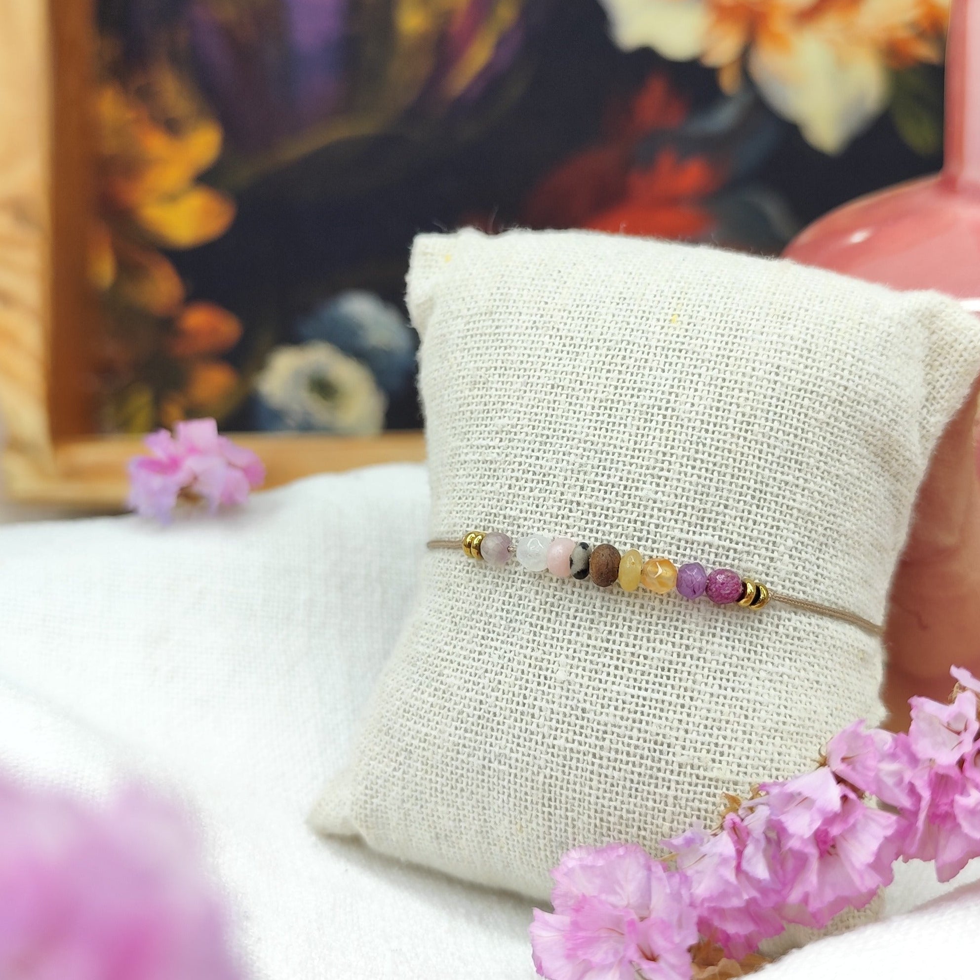 Feines Armband mit Nussperle und bunten Natursteinperlen auf hellem Mischgewebeband, präsentiert auf einem kleinen Stoffkissen, im Vordergrund rosa Blüten