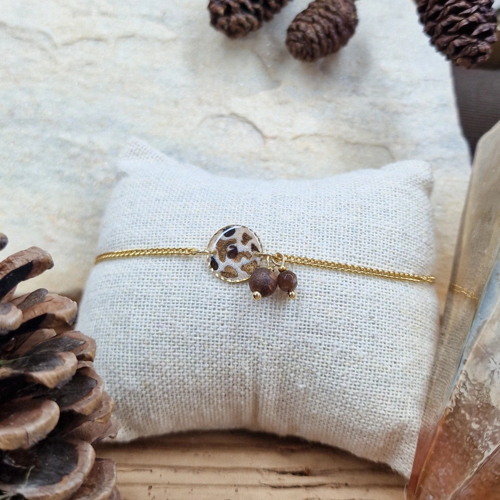Handgefertigtes Armband ‚Leopardis‘ aus Nussholz, hellem Resin-Element im Leo-Design und Natursteinen mit goldfarbenen Edelstahldetails, liegend auf Stoffkissen