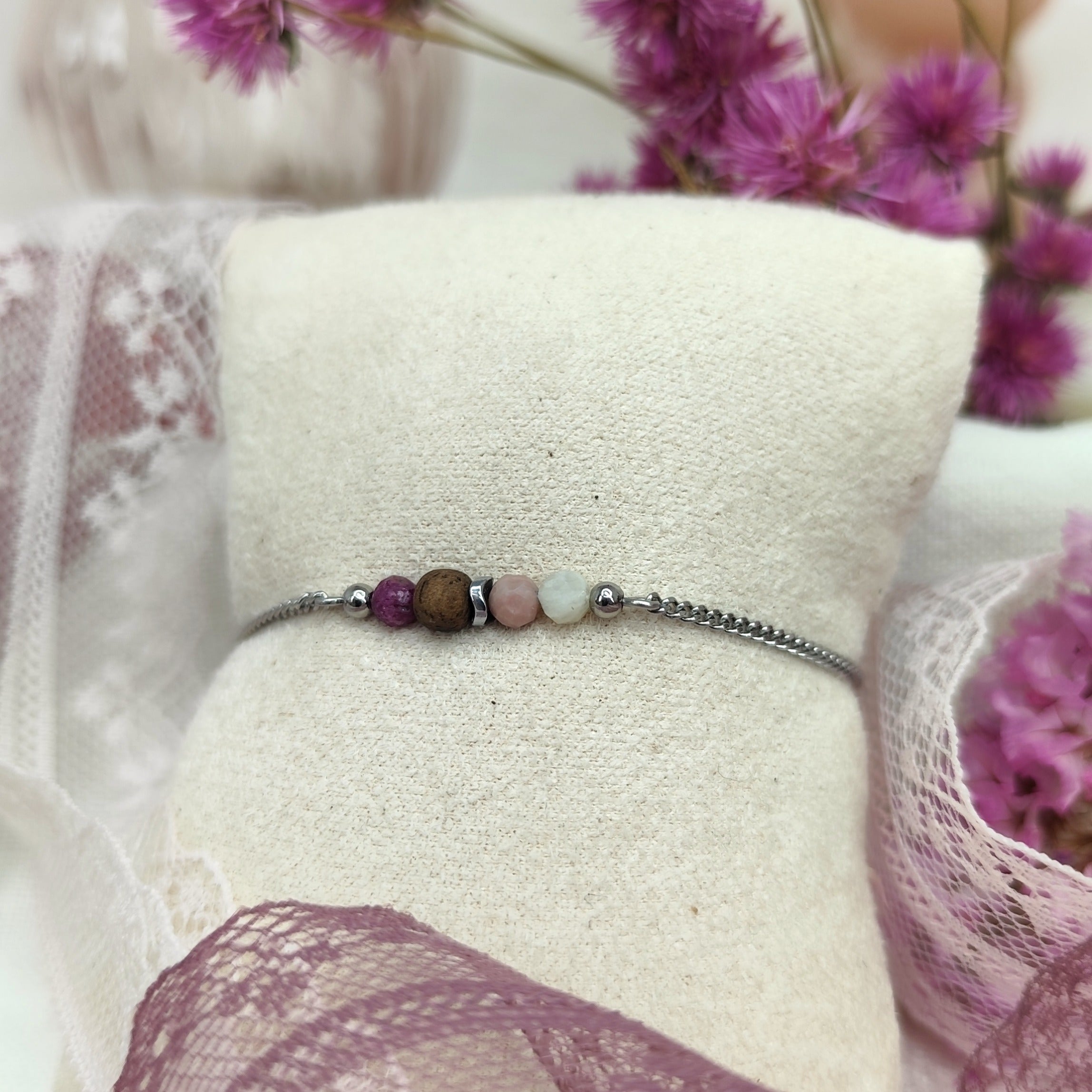 Armketterl mit Nussholzperle und rosé- und violettfarbenen Natursteinen an feiner silberfarbener Kette, auf hellem Stoffpolster und rosa Hintergrund. Spitze und Blumen im Hintergrund