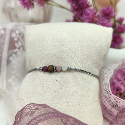 Armketterl mit Nussholzperle und rosé- und violettfarbenen Natursteinen an feiner silberfarbener Kette, auf hellem Stoffpolster und rosa Hintergrund. Spitze und Blumen im Hintergrund