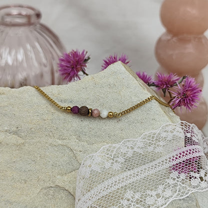 Armketterl mit handgefertigter Nussholzperle, Natursteinen in Rosé- und Violetttönen, arrangiert auf hellem Stein mit rosa Blüten im Hintergrund