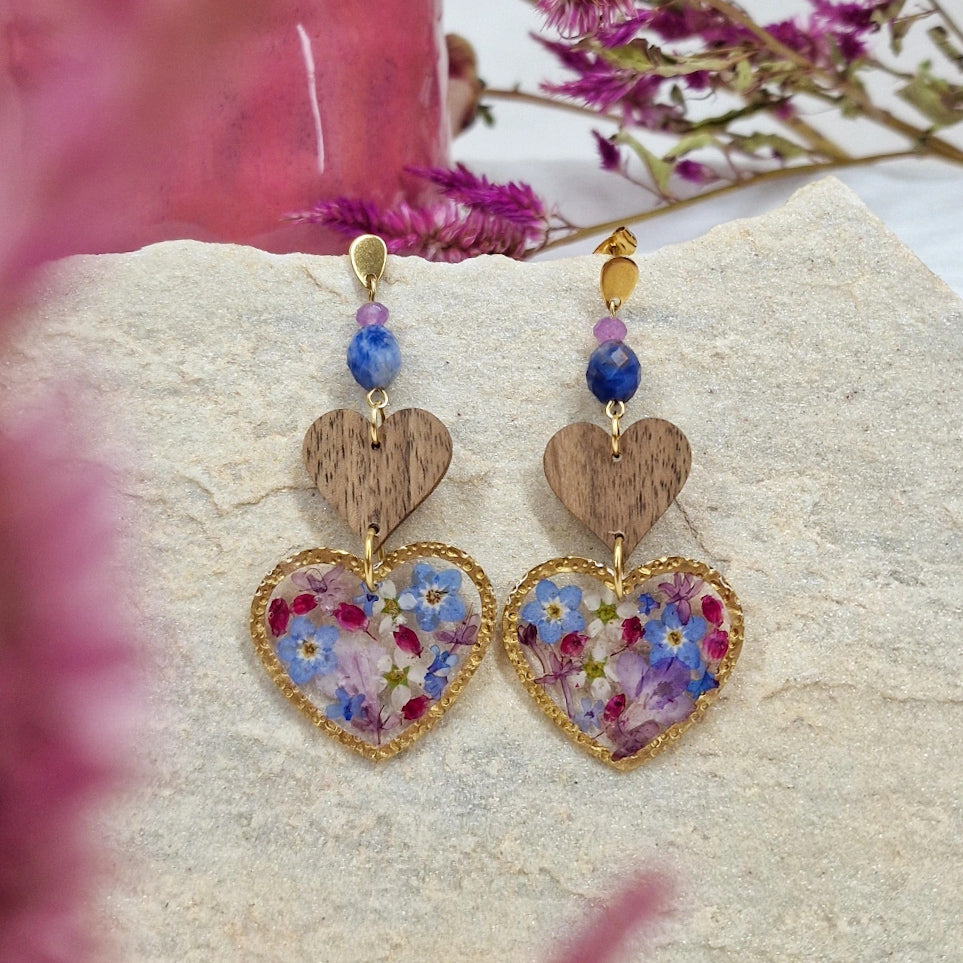Ohrringe mit herzförmigen Resin-Elementen mit echten Blüten und selbstgemachte Herzen aus regionalem Holz und Natursteinen. Arrangiert auf einem hellen Stein mit rosa Hintergrund