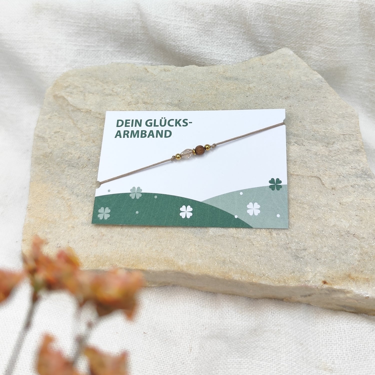 Handgefertigtes Glücksarmband aus Nussholzkugel, tropfenförmig geschliffenem Glasstein und Hämatit auf naturfarbenem Baumwollband, liegend auf hellem Stein neben einer Karte