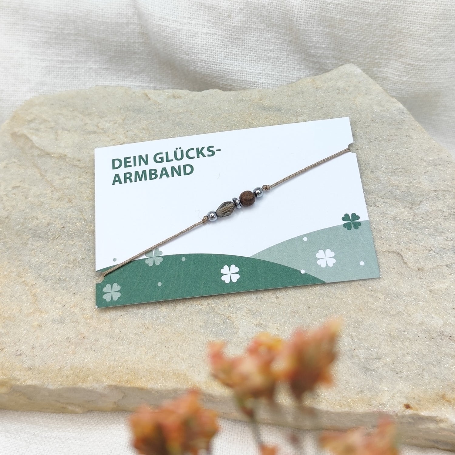 Handgefertigtes Glücksarmband aus Nussholzkugel, tropfenförmig geschliffenem Glasstein und Hämatit auf naturfarbenem Baumwollband, liegend auf hellem Stein neben einer Karte