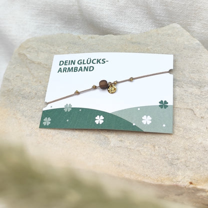 Handgefertigtes Glücksarmband aus Nussholzperle und gehämmertem Edelstahlplättchen gold auf naturfarbenem Baumwollband, liegend auf hellem Stein mit Karte