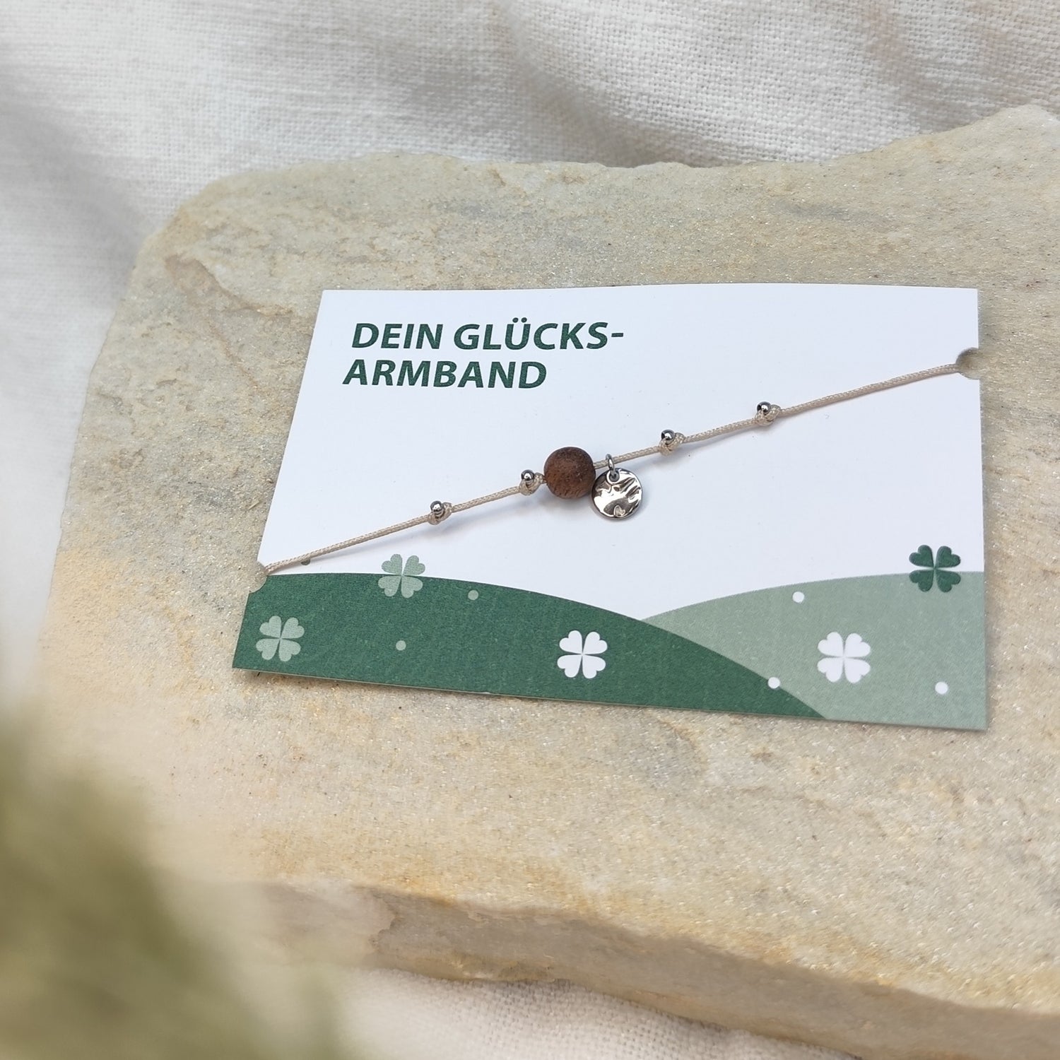 Handgefertigtes Glücksarmband aus Nussholzperle und gehämmertem Edelstahlplättchen silber auf naturfarbenem Baumwollband, liegend auf hellem Stein mit Karte
