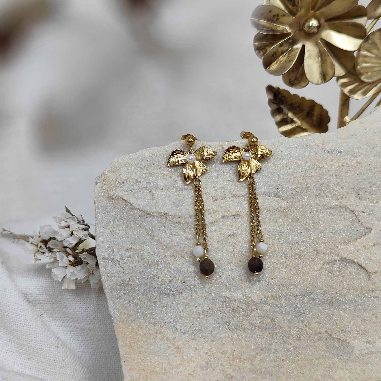 Zwei lange goldfarbene Ohrringe mit Lilienblüte als Stecker, daran ein feines Kettchen mit weißem Naturstein und dunkler Nussperle, präsentiert auf hellem Naturstein