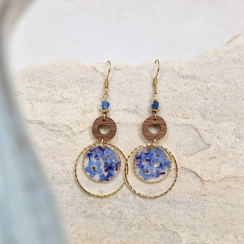 Handgefertigte Ohrringe mit Nussholz, rundem Resin-Element mit echten blauen Vergissmeinnicht-Blüten und Lapislazuli-Detail, arrangiert auf hellem Naturstein.