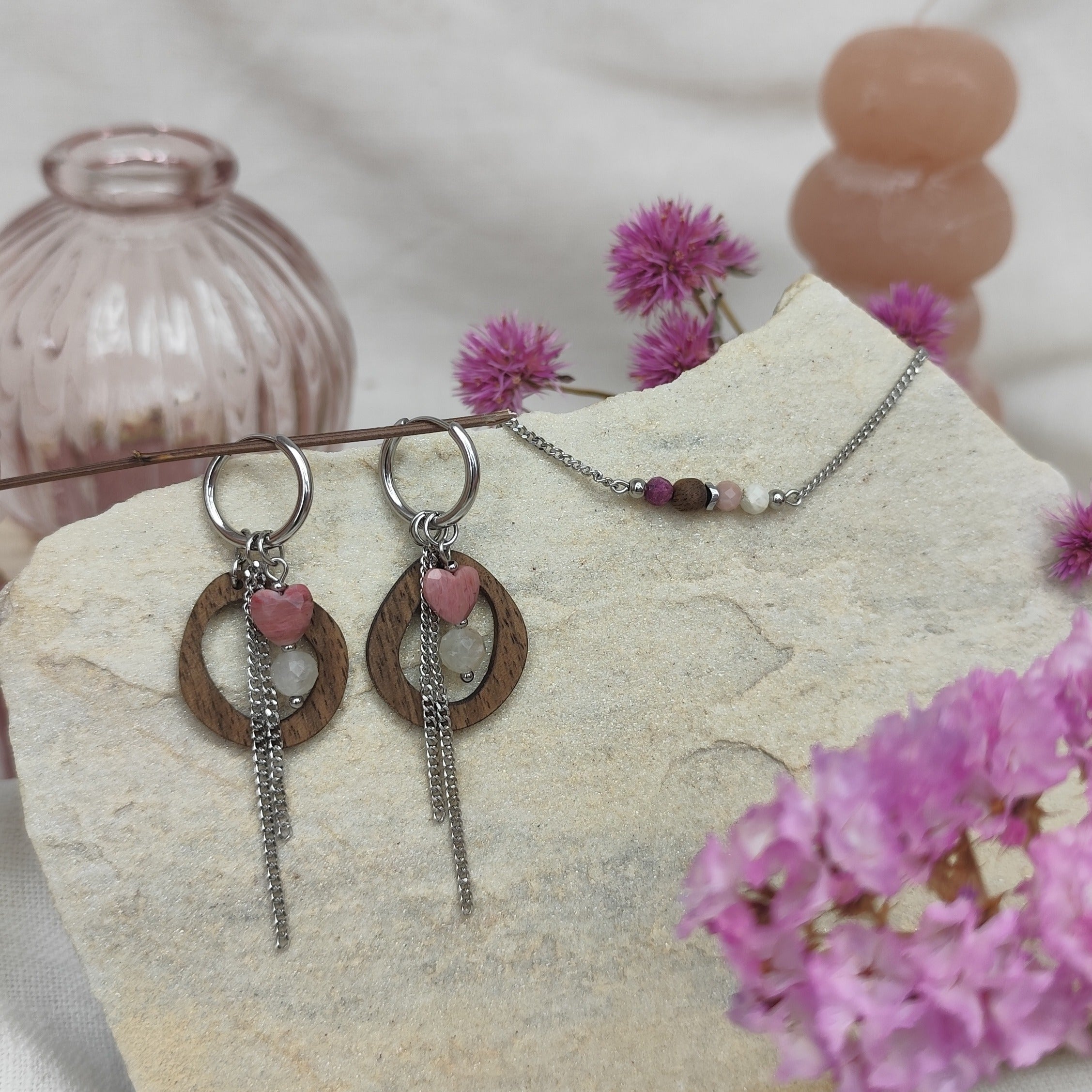 Handgefertigtes Schmuckset mit Mix-and-Match-Kreolen aus Holz und silberfarbenem Metall sowie Armketterl mit kleinen Natursteinen, arrangiert auf hellem Stein und Rosa Blumen in Vorder- und Hintergrund